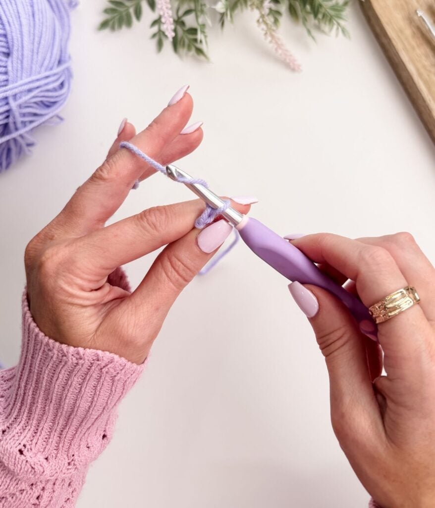 Close-up of hands crocheting a slip knot and chain stitch with a lavender crochet hook and light purple yarn, against a white background with some greenery visible.