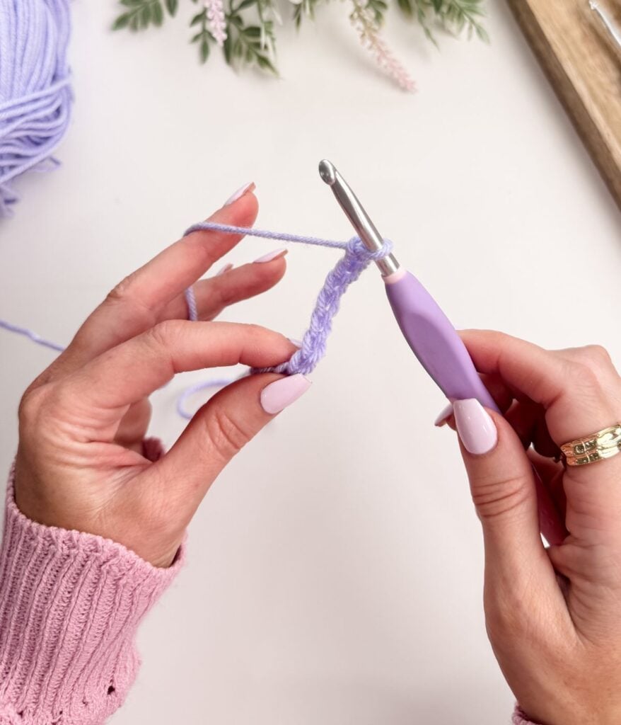 A person with manicured nails holds a crochet hook and lilac yarn, starting with a slip knot and forming a chain stitch on a white surface.