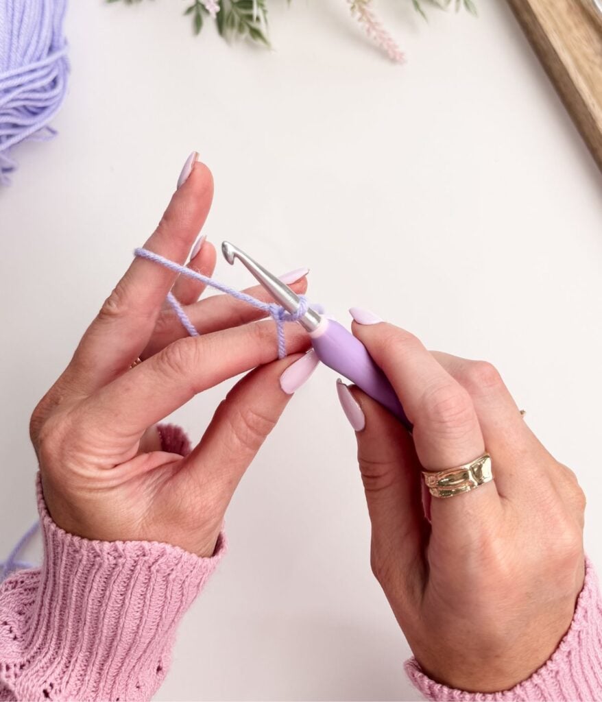 Close-up of hands holding a crochet hook and light purple yarn, preparing to make a slip knot and chain stitch, with a ball of yarn and flowers in the background.