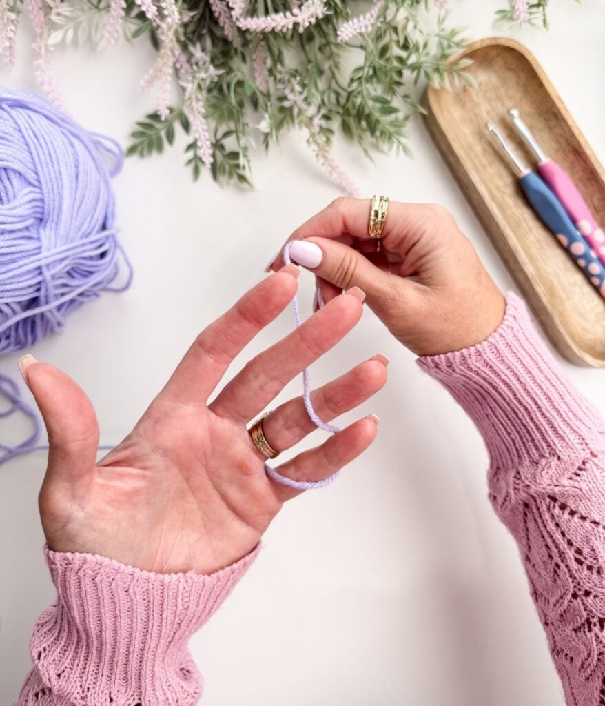 A person wearing a pink sweater wraps light purple yarn around their fingers, with crochet hooks and flowers nearby on a white surface.