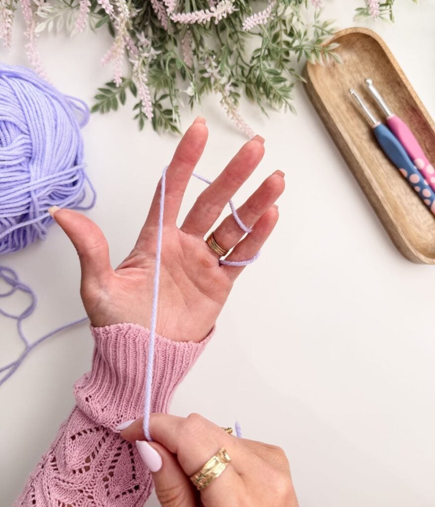 A hand wraps light purple yarn around fingers, with crochet hooks and a wooden tray nearby on a white surface.