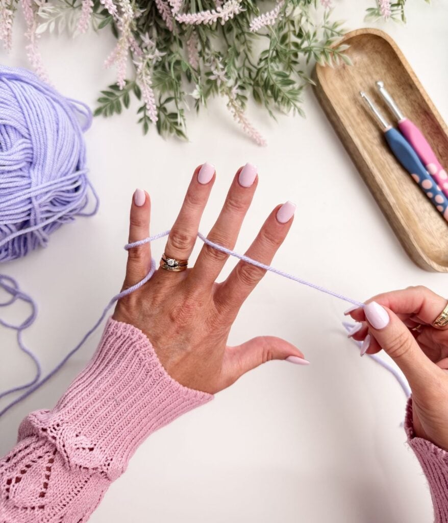 A person wearing a pink sweater wraps lavender yarn around their fingers, with crochet hooks and a ball of yarn on a white surface.