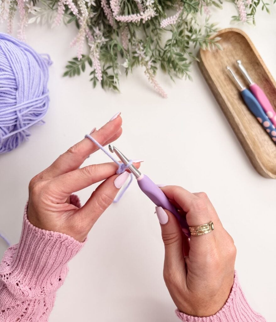 A person in a pink sweater crochets with lavender yarn using a crochet hook, with extra hooks and flowers on a white table.