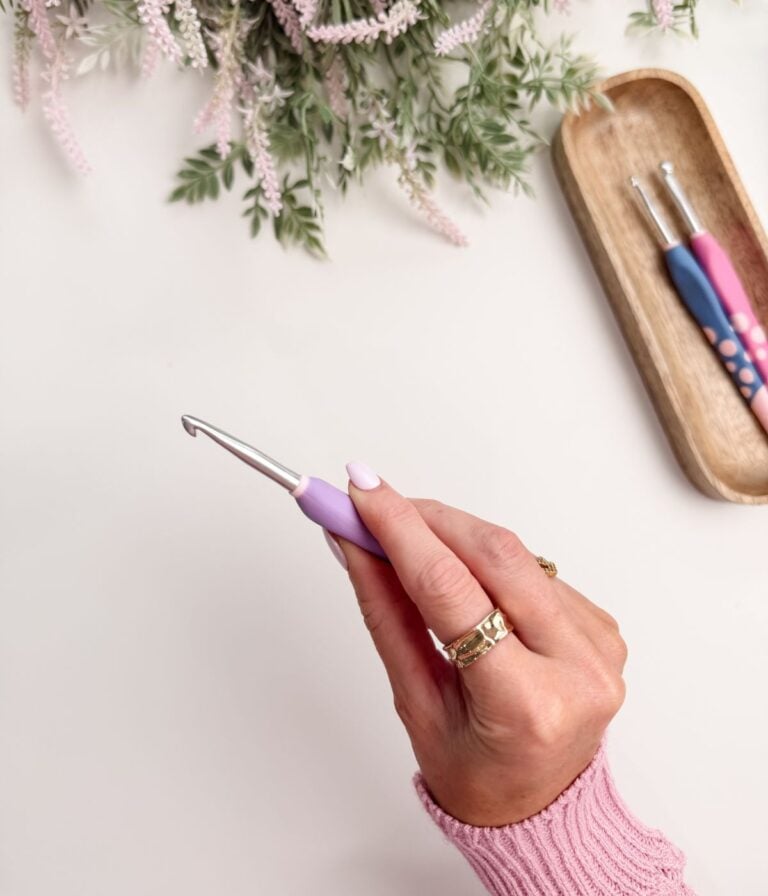 A hand holding a purple crochet hook, with a tray holding two more crochet hooks and some greenery in the background.