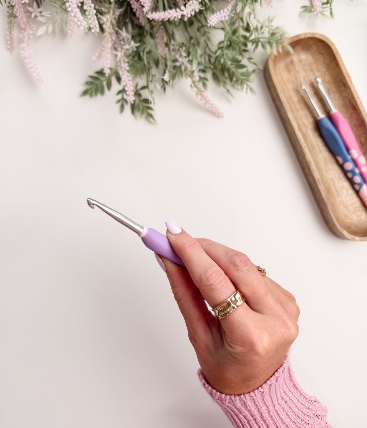 A hand holding a purple crochet hook, with a tray holding two more crochet hooks and some greenery in the background.