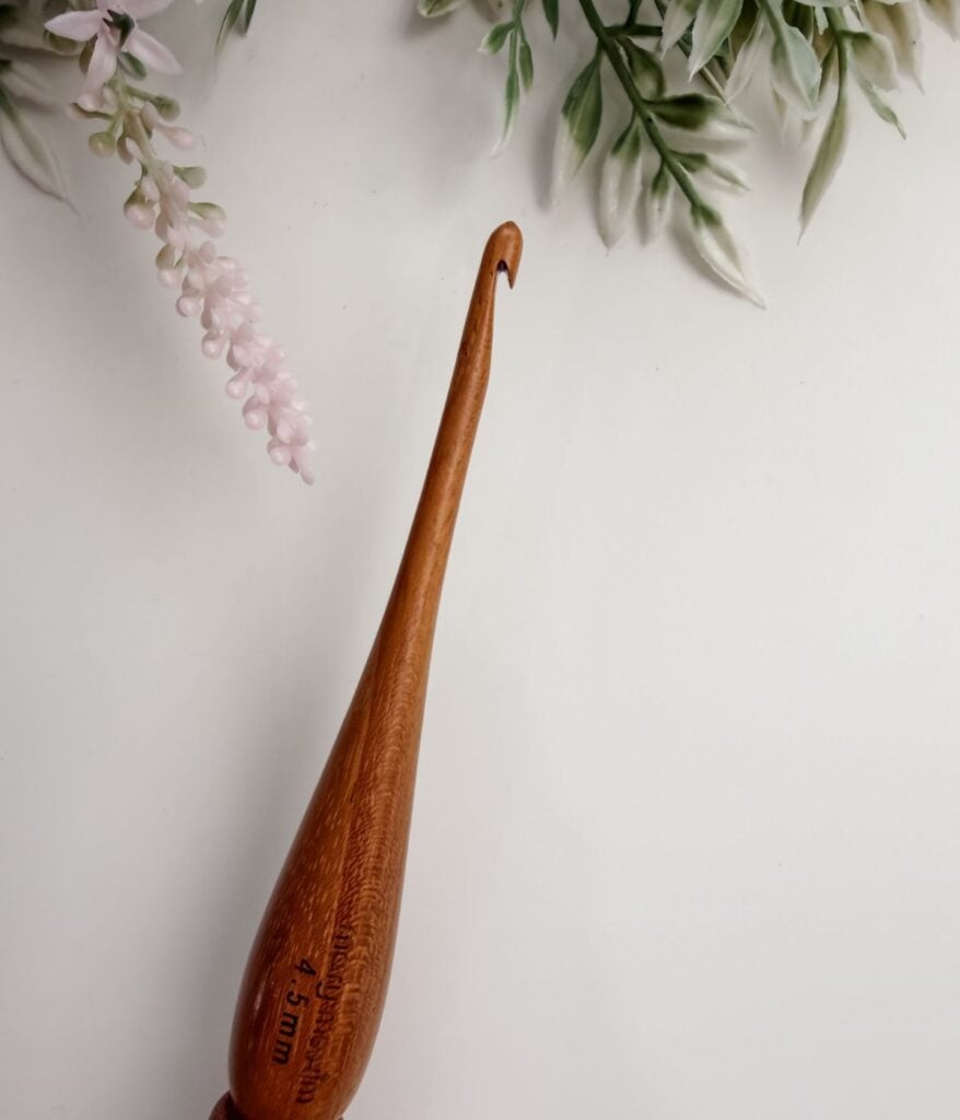 A wooden crochet hook with a 3.5mm size marking, ideal for those learning about crochet hook sizes and materials, rests on a white surface with green and pink foliage in the background.
