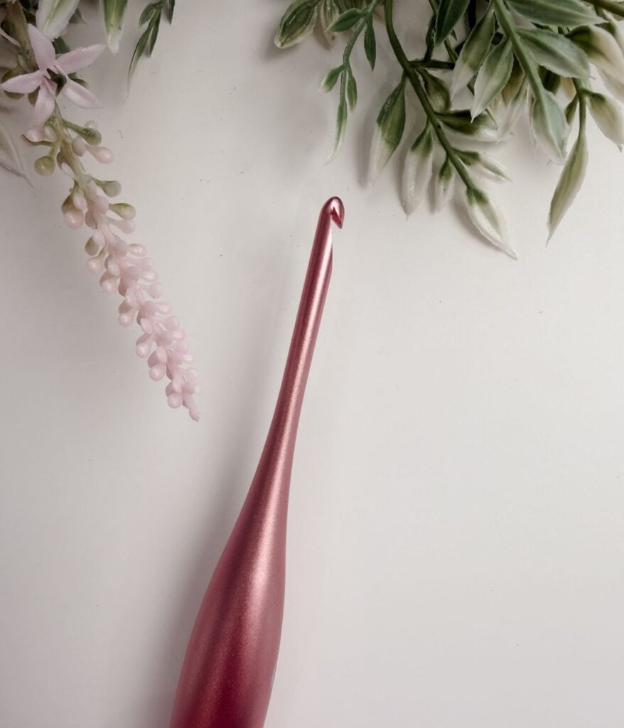 A metallic pink crochet hook, ideal for beginners exploring different crochet hook sizes and materials, is positioned on a white surface with artificial green and pink foliage in the corners.