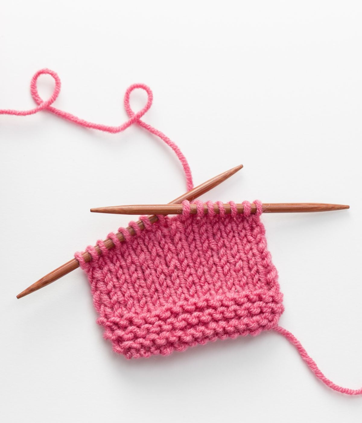 Two wooden knitting needles hold a partially knitted piece of pink yarn on a white background, with the yarn trailing off in loops.