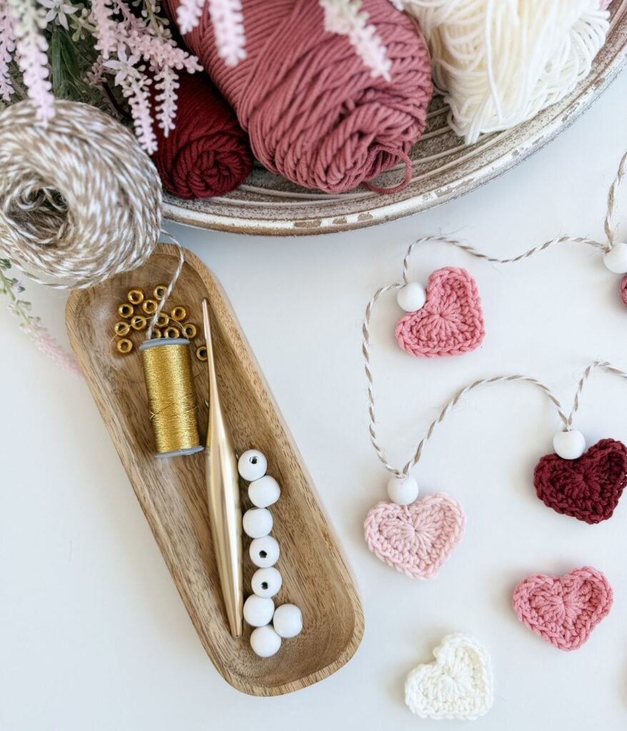 A bowl of yarn, a tray with gold thread, beads, and a crochet needle, and a heart garland of crocheted hearts and white beads are arranged on a white surface.