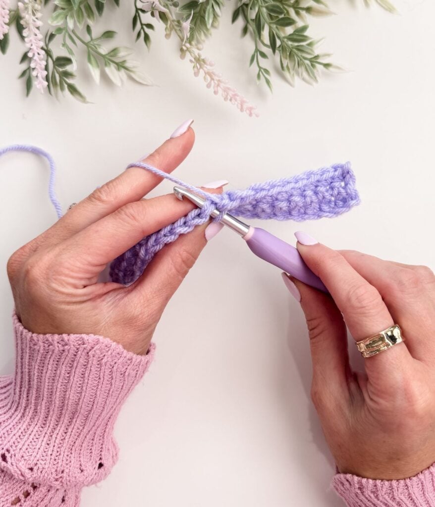 Hands crocheting with a purple hook and lavender yarn on a white surface, demonstrating single crochet for beginners, with green and pink foliage in the background.