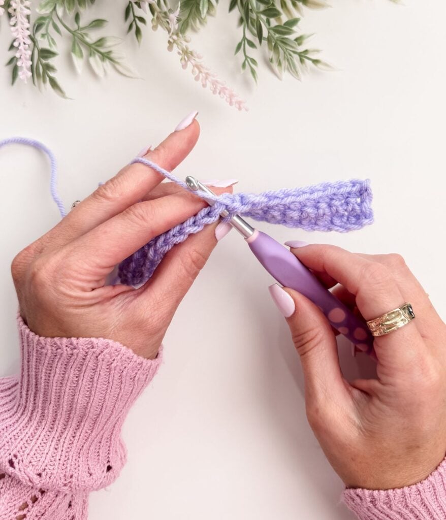 Hands demonstrate single crochet for beginners with a piece of purple yarn and matching hook; person wears a pink sweater and has manicured nails. Greenery accents the top of the image.