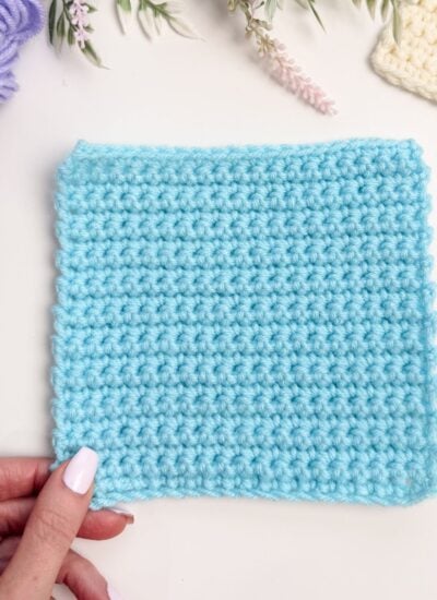 A hand holding a square piece of light blue crochet fabric, perfect for practicing single crochet for beginners, displayed on a white surface with yarn and crocheted items in the background.