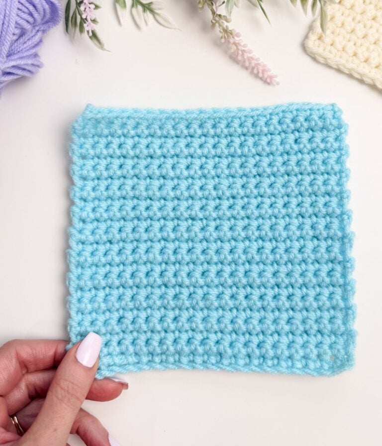 A hand holding a square piece of light blue crochet fabric, perfect for practicing single crochet for beginners, displayed on a white surface with yarn and crocheted items in the background.