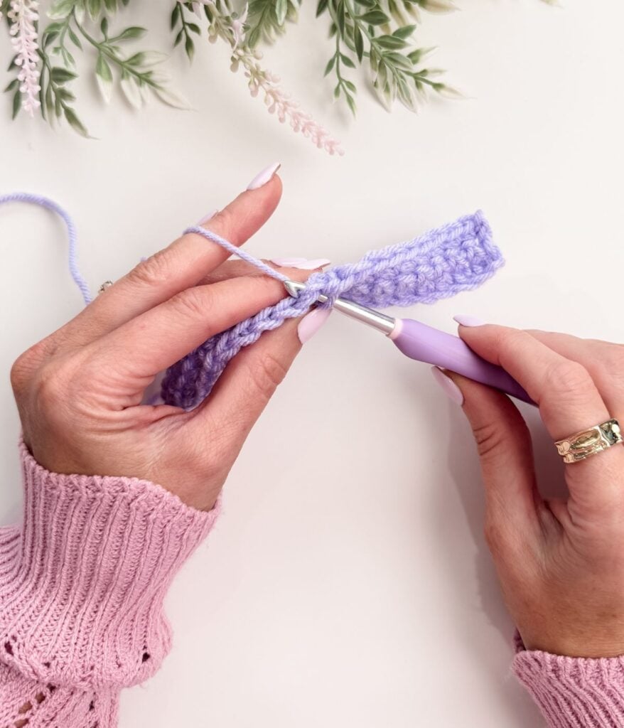 Two hands crocheting with a lavender yarn and hook using the single crochet for beginners technique; person wears a pink sweater and gold ring, with greenery visible at the top of the image.