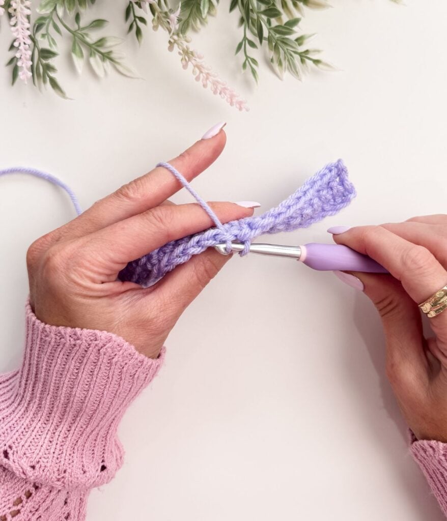 Hands crocheting a row with light purple yarn and a purple crochet hook, perfect for practicing single crochet for beginners, with leafy sprigs in the background on a white surface.