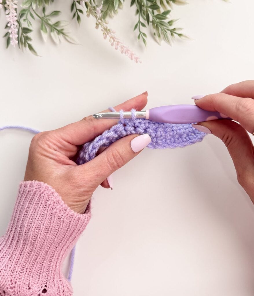 Close-up of hands demonstrating single crochet for beginners with lavender yarn and a purple crochet hook, wearing a pink knitted sweater, with green leaves in the background.