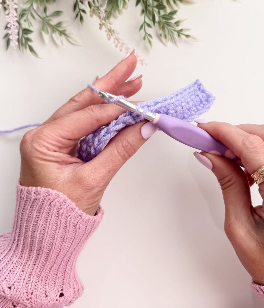 Hands crocheting a lavender yarn using a purple-handled hook, demonstrating single crochet for beginners, with green foliage in the background.