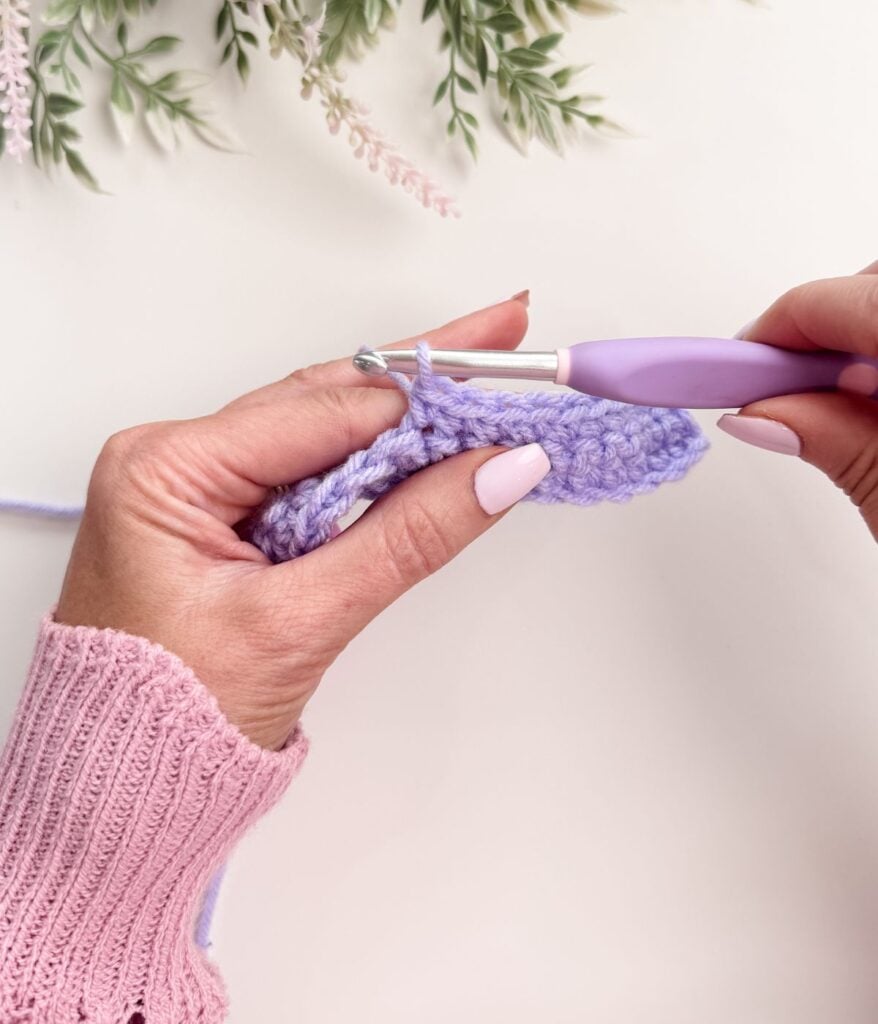 A person wearing a pink sweater is crocheting with a purple crochet hook and light purple yarn, demonstrating single crochet for beginners on a white surface with green foliage in the background.
