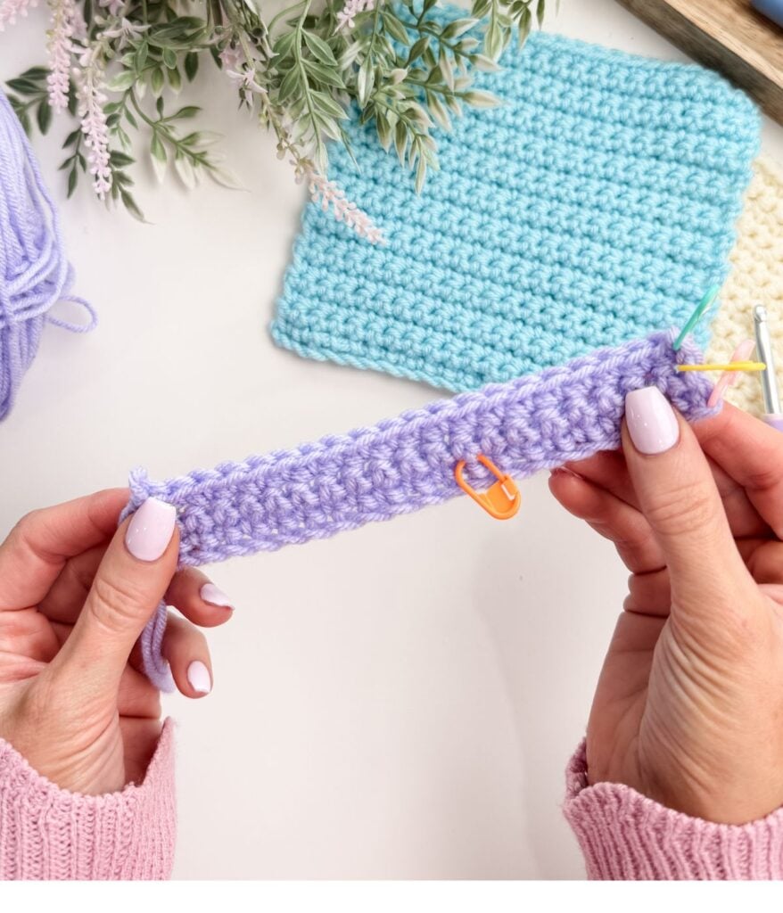 Hands holding a piece of purple crochet with a stitch marker, yarn, and a finished blue crochet square on the table—perfect for practicing single crochet for beginners.