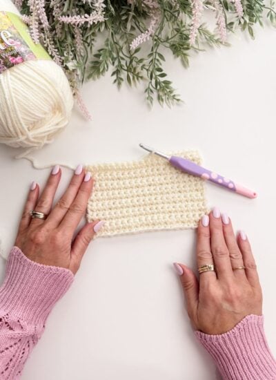 Hands with pale pink nail polish hold a crocheted swatch next to a skein of white yarn and a crochet hook, with flowers partially visible at the top.