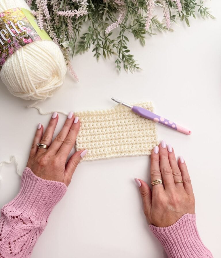 Hands with pale pink nail polish hold a crocheted swatch next to a skein of white yarn and a crochet hook, with flowers partially visible at the top.