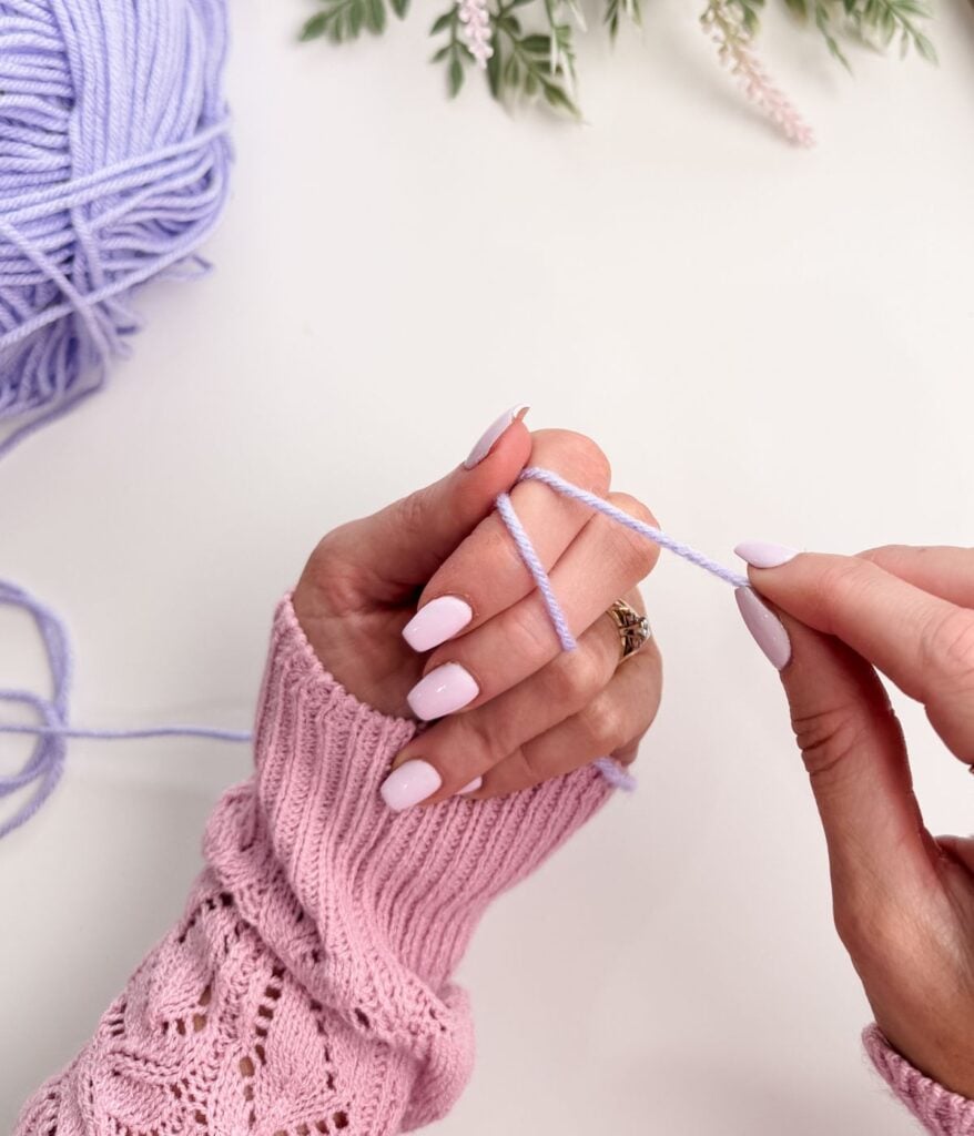 A person wearing a pink sweater starts a crochet project with light purple yarn, forming a slip knot and chain stitch as they wrap the yarn around their fingers.
