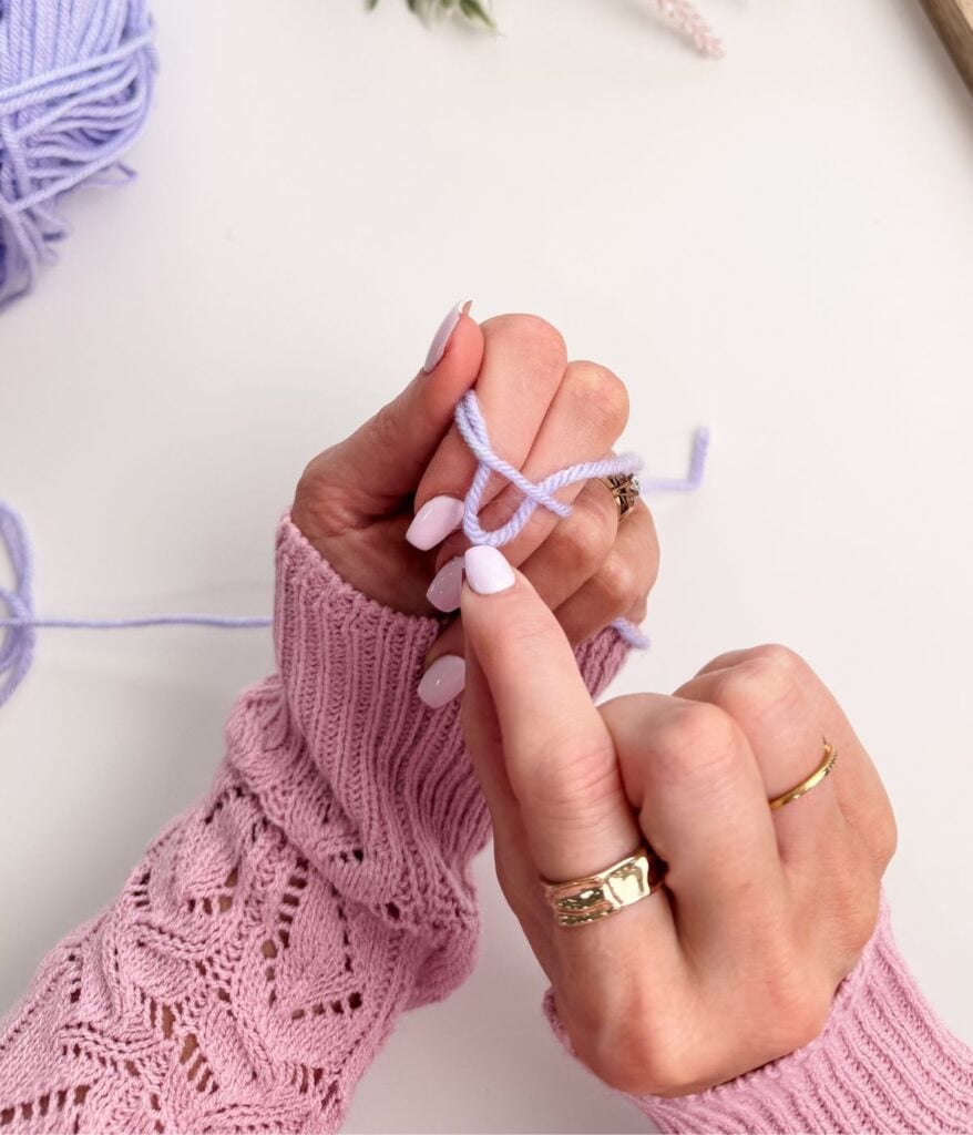 A person wearing a pink knit sweater forms a slip knot and casts on light purple yarn with their fingers, preparing to knit a chain stitch.