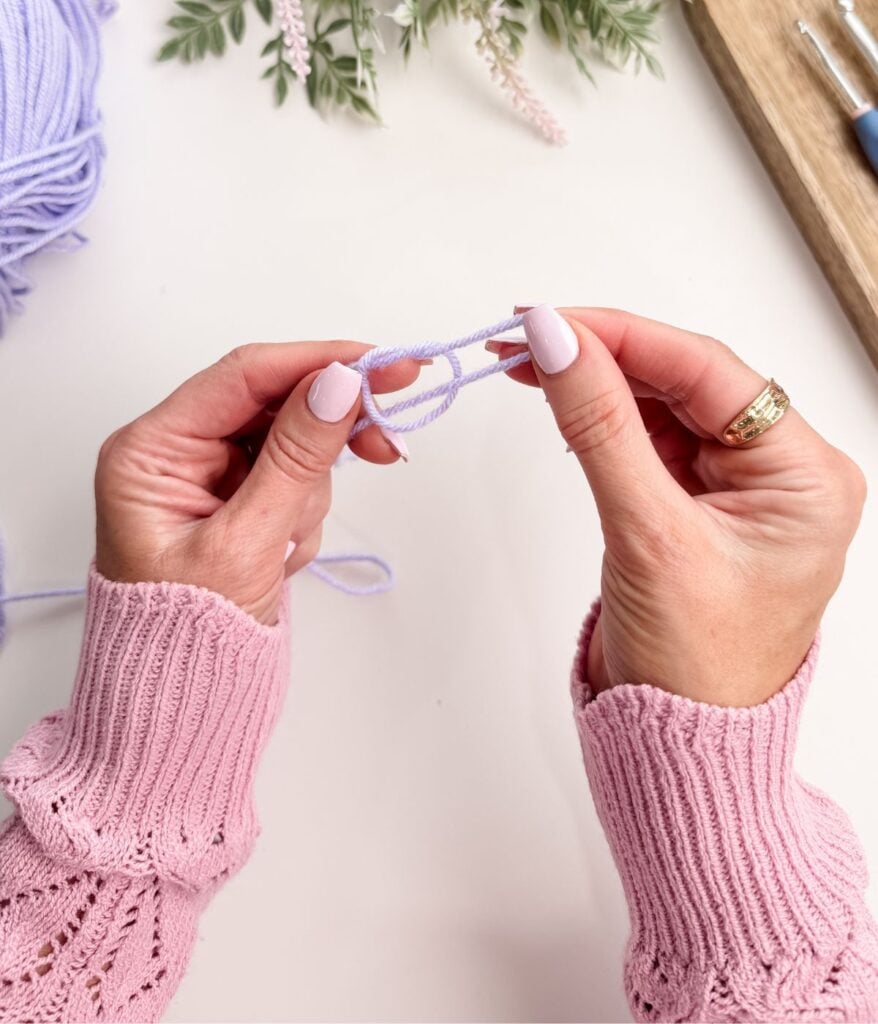 Two hands with light pink nail polish hold a loop of light purple yarn, ready to create a slip knot or begin a chain stitch; crochet tools and yarn are visible on a white surface.
