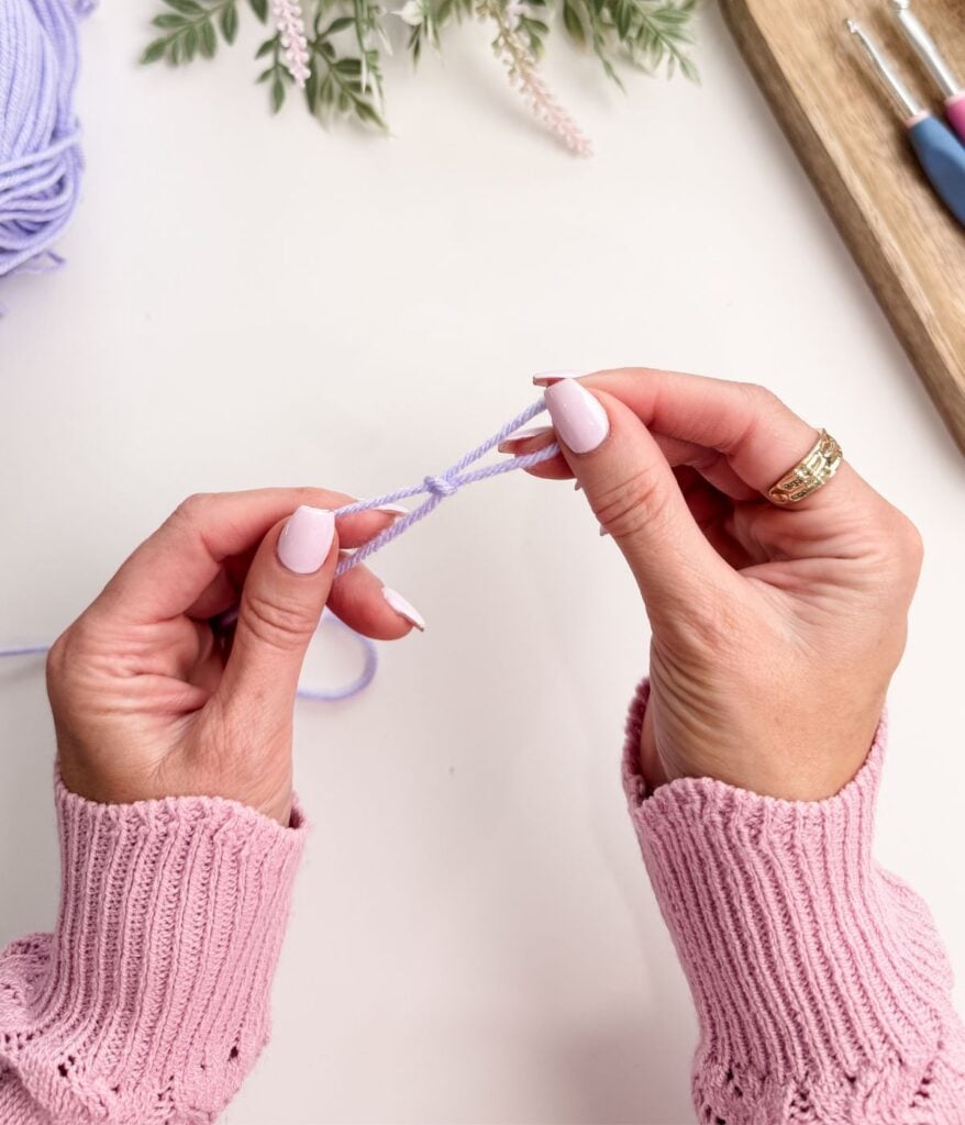 Close-up of hands in a pink sweater holding and twisting light purple yarn, preparing a slip knot and chain stitch, with crochet hooks and flowers in the background.