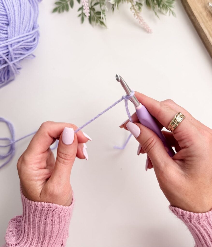 Close-up of hands making a slip knot and chain stitch with light purple yarn, starting a crochet project on a white surface.