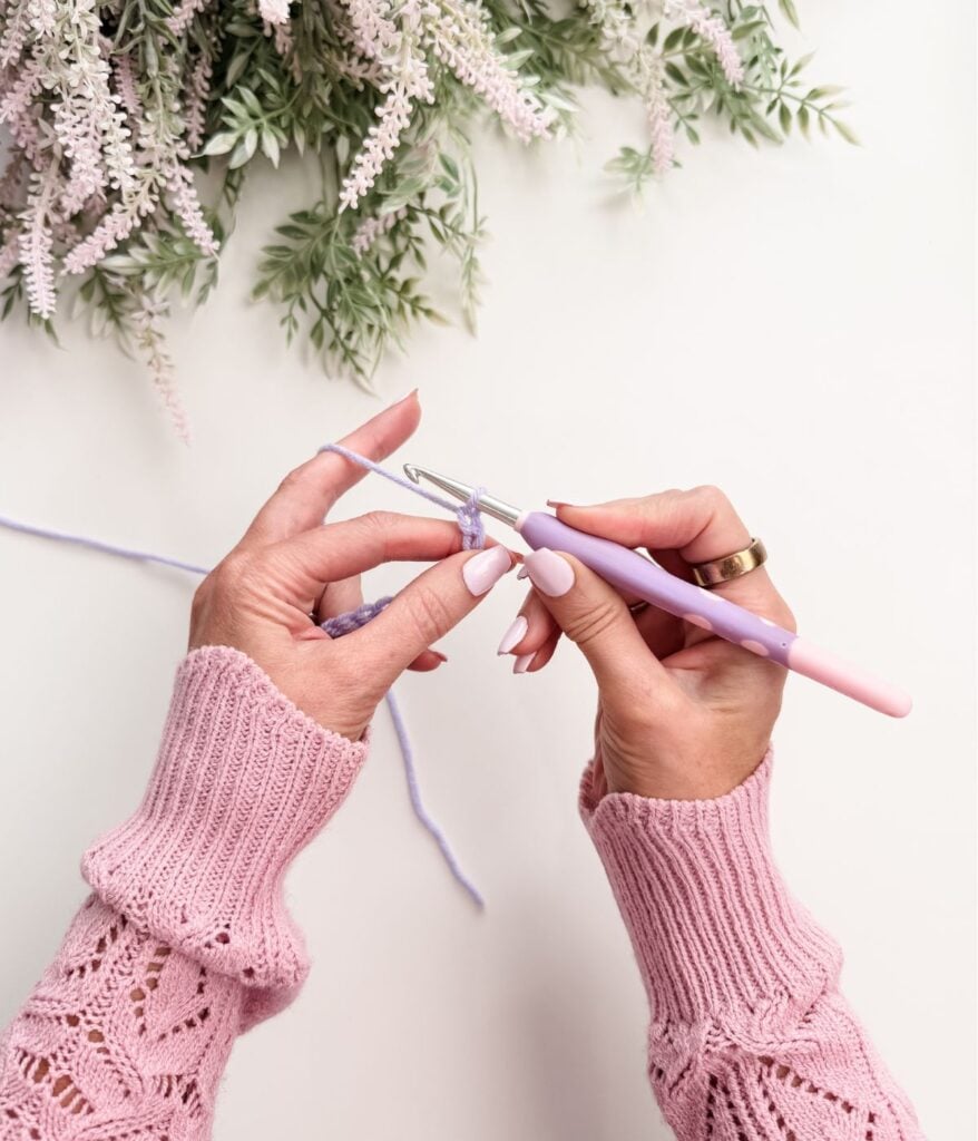 A person wearing a pink sweater is crocheting with a lavender yarn and a pink crochet hook, demonstrating how to hold a crochet hook; flowers are visible at the top of the image.