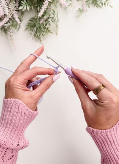Hands demonstrating how to hold a crochet hook for beginners with a purple hook and lavender yarn, wearing a pink sweater and gold ring, surrounded by greenery in the background.
