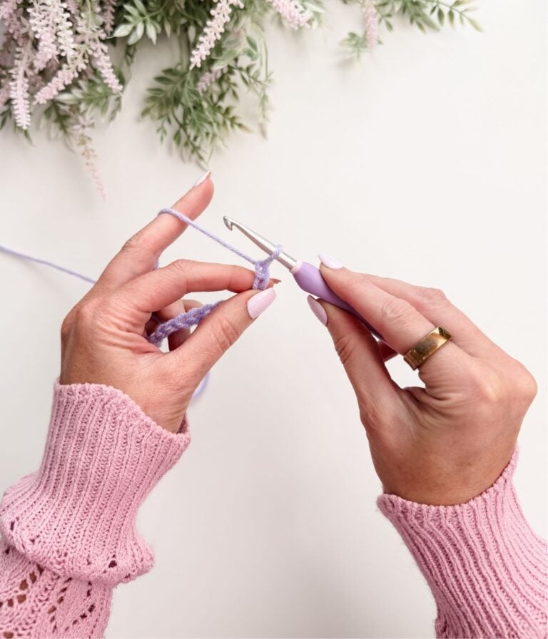 Hands demonstrating how to hold a crochet hook for beginners with a purple hook and lavender yarn, wearing a pink sweater and gold ring, surrounded by greenery in the background.