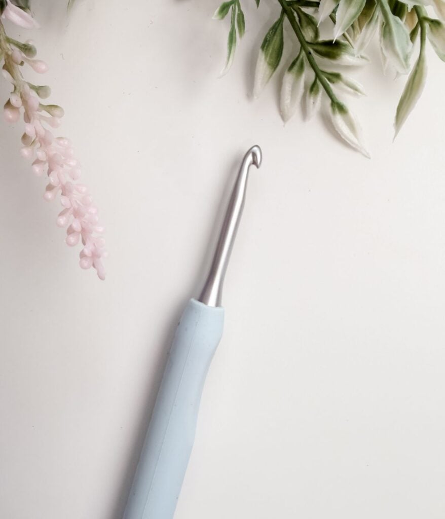A metal crochet hook with a light blue handle rests on a white surface, surrounded by green and pink artificial foliage—an ideal choice for those seeking the best crochet hooks for beginners.