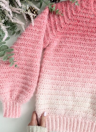 A hand holds the bottom of a pink and white ombre crocheted sweater placed on a flat surface next to some greenery and lavender sprigs.