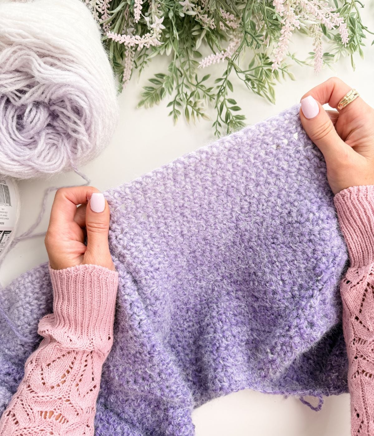 Hands holding a partially knitted purple fabric with a ball of matching yarn and greenery in the background.