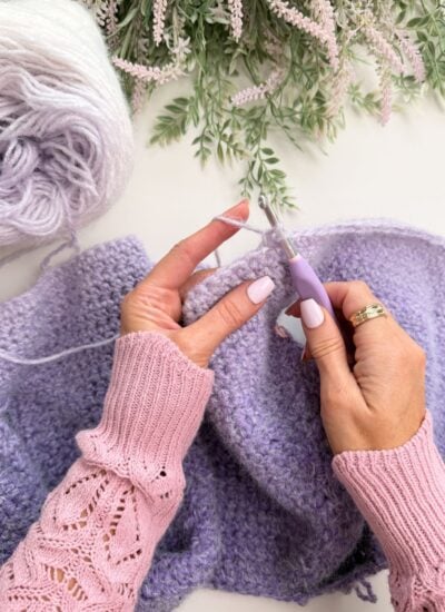 Hands crocheting a lavender-colored yarn piece with a crochet hook, resting on a white surface with a ball of yarn and greenery nearby.