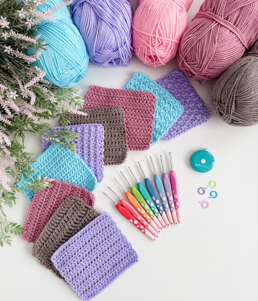 Yarn balls, crocheted swatches, colorful crochet hooks, stitch markers, and a tape measure arranged on a white surface with decorative greenery.