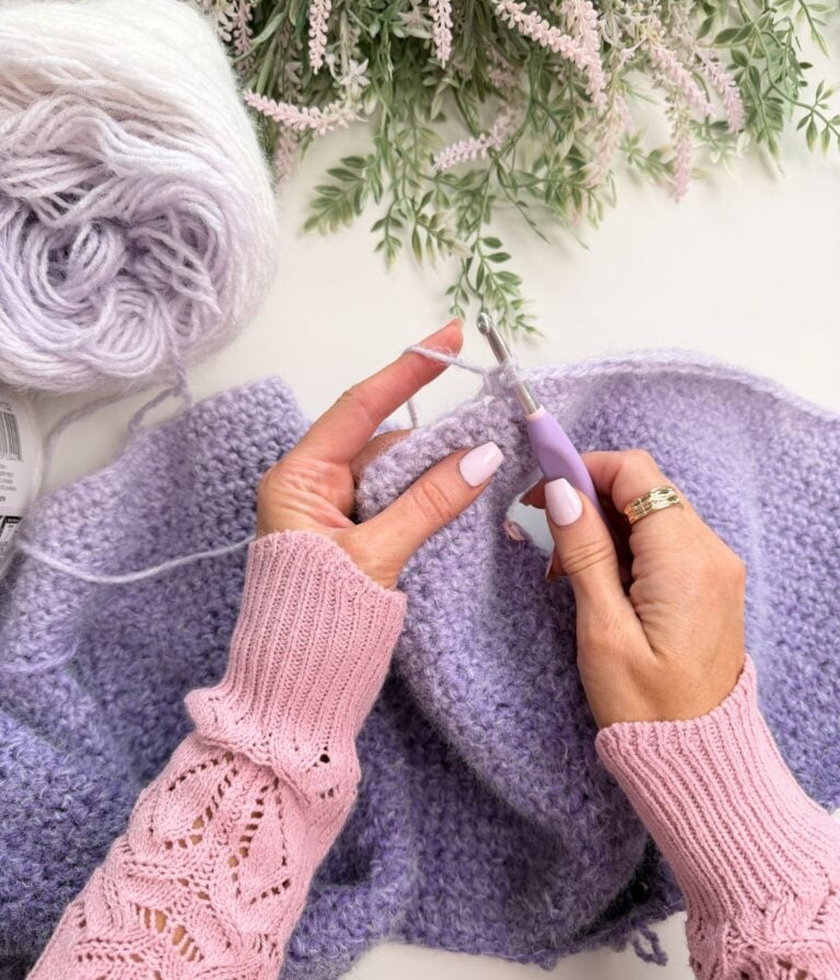 Hands crocheting a lavender-colored yarn piece with a crochet hook, resting on a white surface with a ball of yarn and greenery nearby.