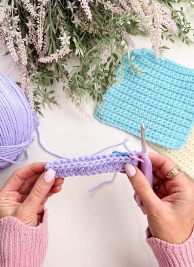 Hands holding a crochet project with lavender yarn, a crochet hook, and finished blue and cream crochet squares on a white surface with green foliage above.