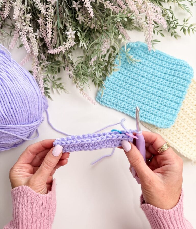 Hands holding a crochet project with lavender yarn, a crochet hook, and finished blue and cream crochet squares on a white surface with green foliage above.