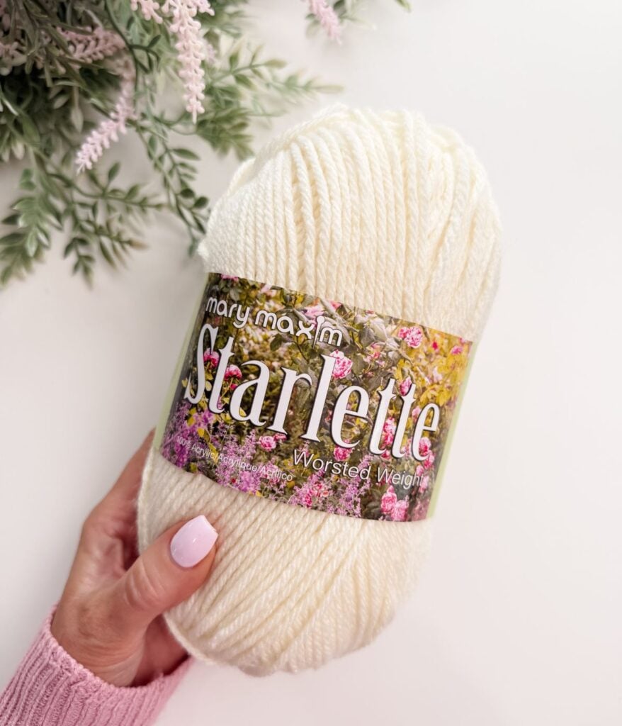 A hand holds a skein of white Mary Maxim Starlette worsted weight yarn, with greenery and a pink sweater sleeve visible.