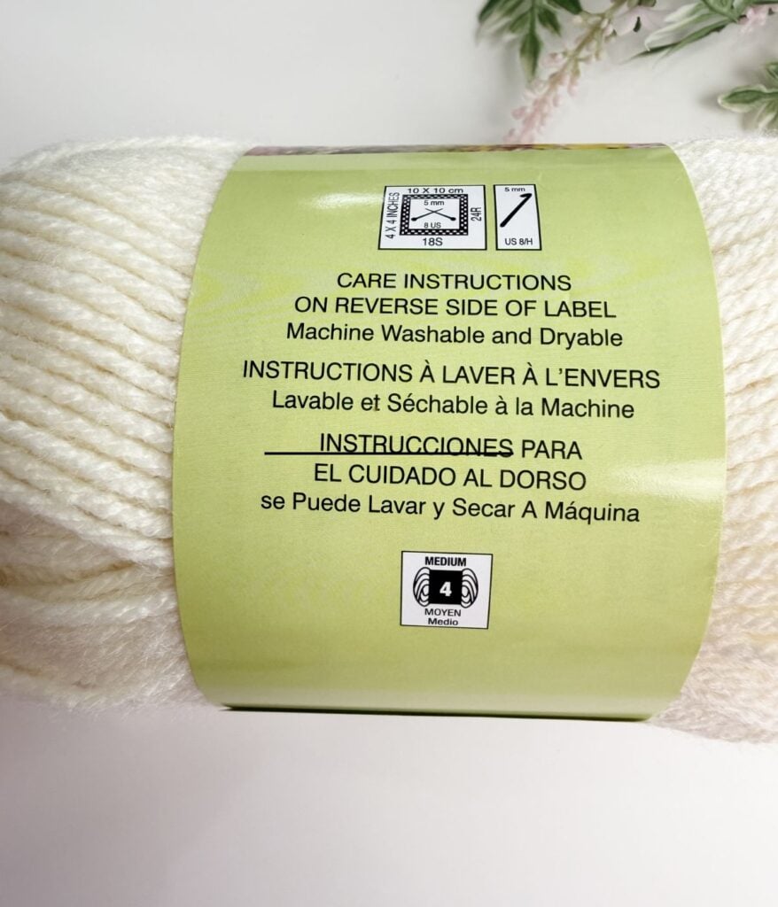 A skein of cream-colored yarn with a green label showing care instructions in English, French, and Spanish for machine washing and drying, and laundry symbols.