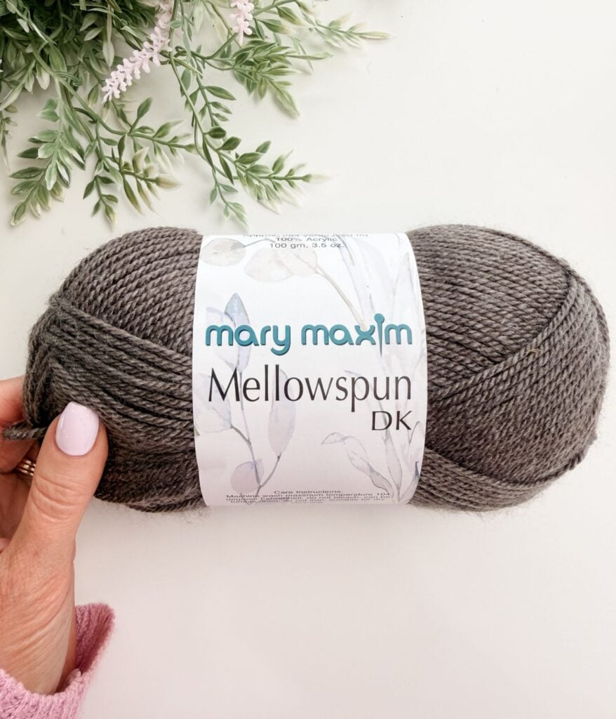 A hand holds a skein of Mary Maxim Mellowspun DK yarn in gray color against a white background with green leafy decor.