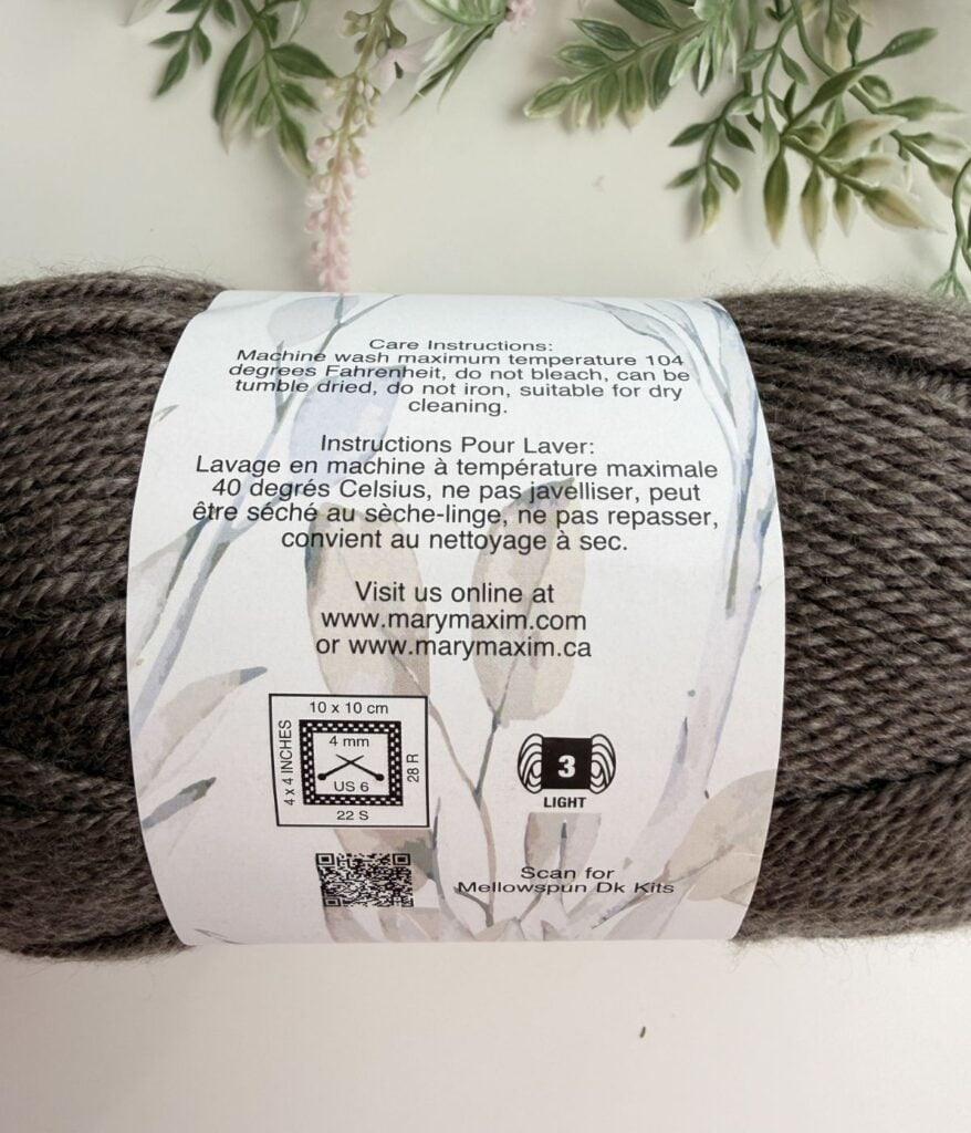 Close-up of a yarn label showing care instructions, washing symbols, website details, and a QR code on a gray skein with leaves in the background.