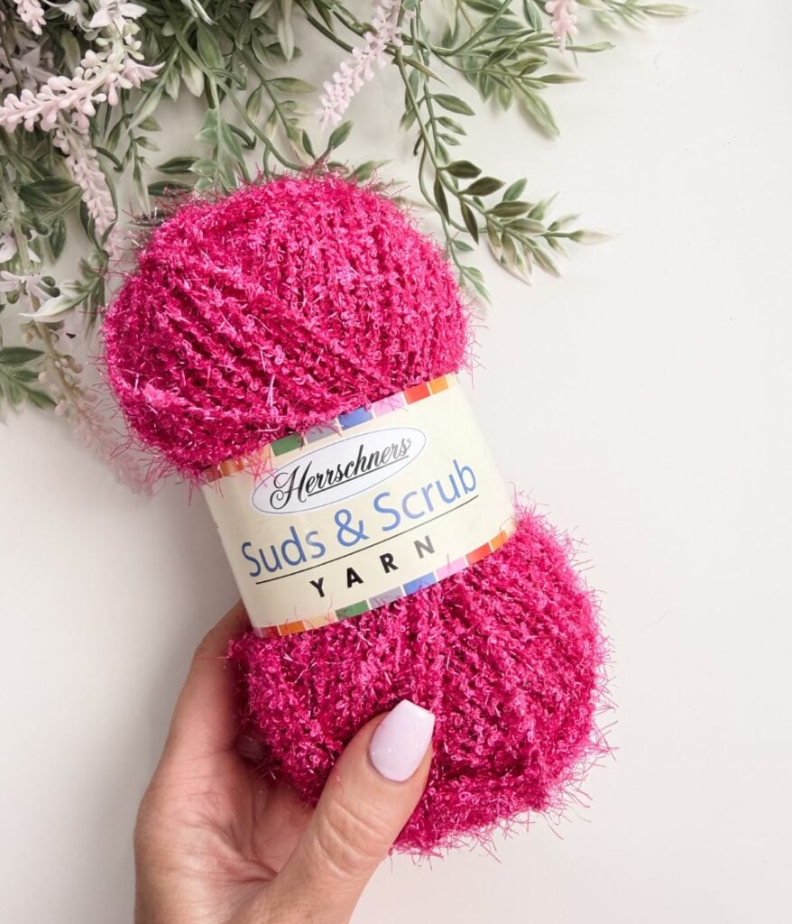 A hand holding a skein of bright pink Herschners Suds & Scrub yarn, with greenery and flowers in the background.