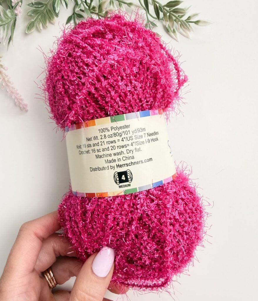 A hand holds a skein of bright pink fuzzy yarn labeled as 100% polyester, medium weight, 2.8 oz, distributed by Herrschners. Greenery decor is in the background.