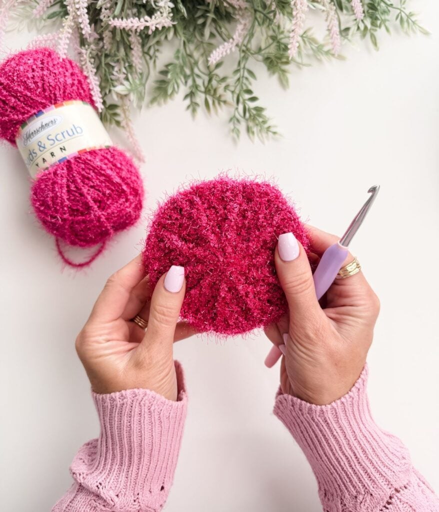 Hands holding a pink crocheted scrubber with a crochet hook, pink yarn and greenery visible on a white surface.