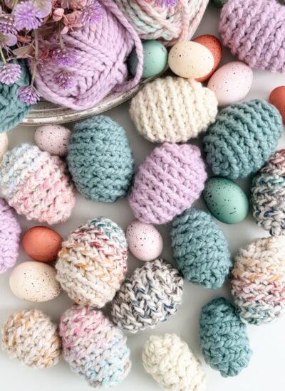 Assorted pastel-colored knitted Easter eggs and speckled decorative eggs are arranged with yarn balls and purple flowers on a white surface.