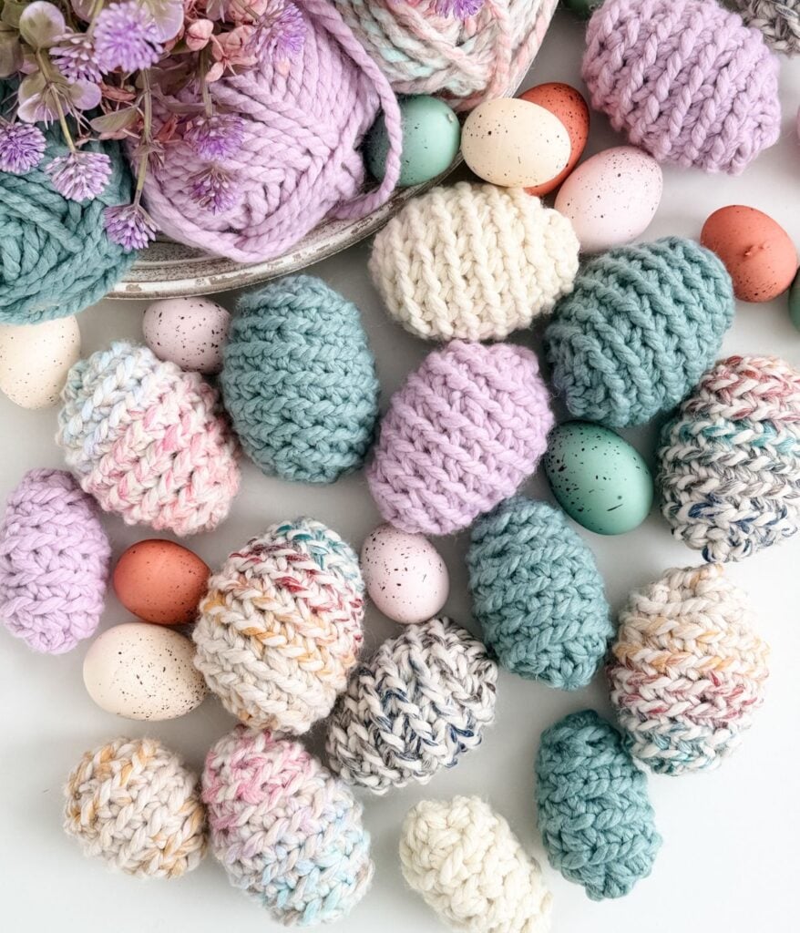 Assorted pastel-colored knitted Easter eggs and speckled decorative eggs are arranged with yarn balls and purple flowers on a white surface.